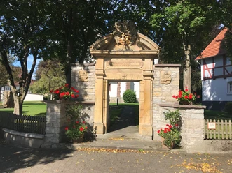 Historischer Torbogen aus Sandstein mit Blumen, flankiert von Bäumen, führt zu einem gepflegten Platz.