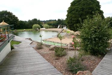 Naturfreibad inmitten grüner Umgebung mit Sandstrand, Rutsche und gepflegten Holzstegen.