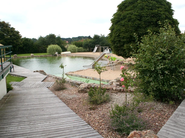 Naturfreibad inmitten grüner Umgebung mit Sandstrand, Rutsche und gepflegten Holzstegen.