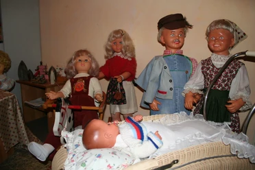 Puppenmuseum Eine Gruppe fröhlicher Puppen in traditioneller Kleidung umgibt einen Kinderwagen mit einem Baby.
