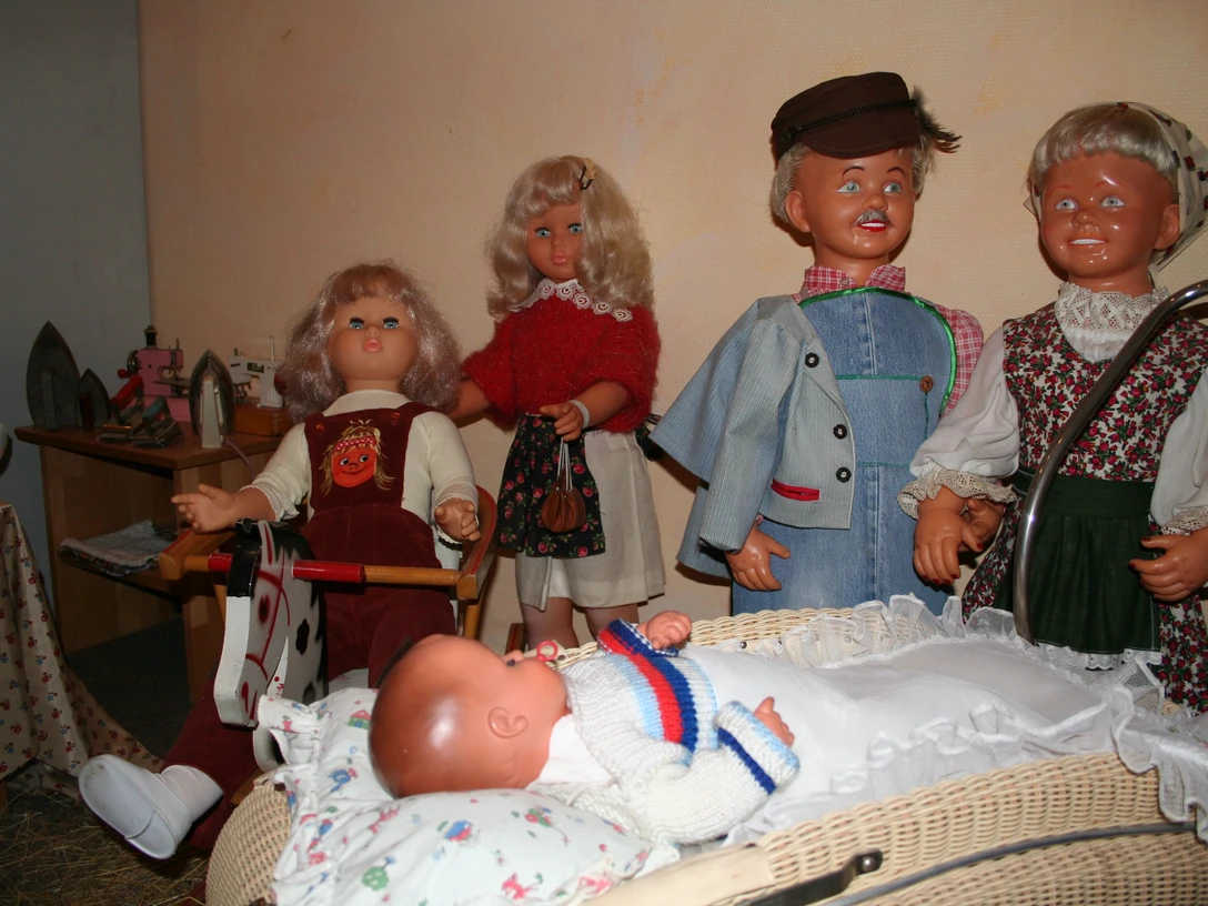 Puppenmuseum Eine Gruppe fröhlicher Puppen in traditioneller Kleidung umgibt einen Kinderwagen mit einem Baby.