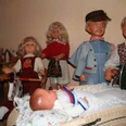 Puppenmuseum Eine Gruppe fröhlicher Puppen in traditioneller Kleidung umgibt einen Kinderwagen mit einem Baby.
