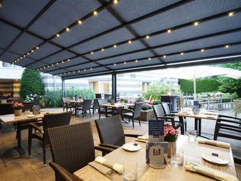 Überdachte Terrasse eines Restaurants mit gedeckten Tischen, Rattanmöbeln und umgebender Begrünung.