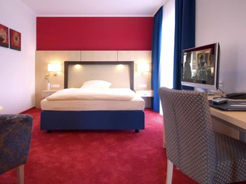 Hotelzimmer Komfort Plus Helles Hotelzimmer mit Doppelbett, Schreibtisch, TV, roten Akzentwänden und einem Fenster mit Vorhängen.
