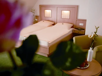 Hotelzimmer Komfort Doppelbett mit hellem Holzkopfteil, grüner Teppich, Sessel und Tisch mit dekorativer Blume.