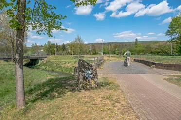 Brücke aus Buntsandstein- Startpunkt Rundweg Brücken und Grenzen Brücke aus rötlichem Sandstein über einen Bach, umgeben von Wiesen und Bäumen, mit Radfahrer.
