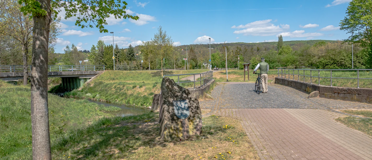 Brücke aus Buntsandstein- Startpunkt Rundweg Brücken und Grenzen Brücke aus rötlichem Sandstein über einen Bach, umgeben von Wiesen und Bäumen, mit Radfahrer.