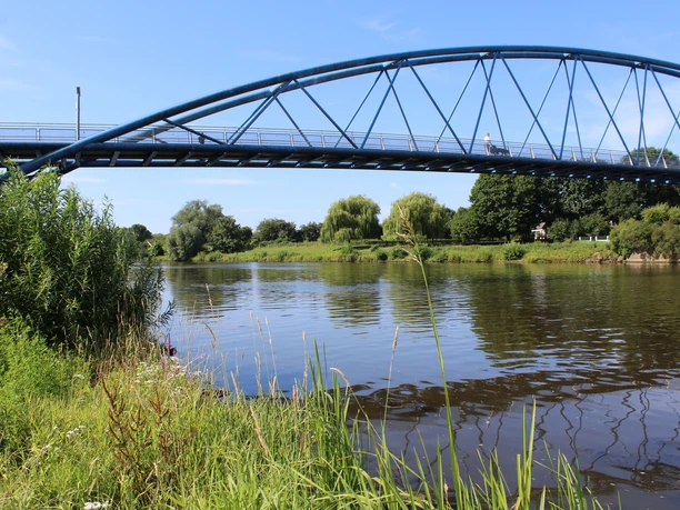 Blue arch bridge spans a river, surrounded by green vegetation and a clear summer sky.