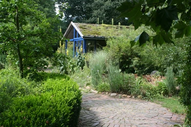 Versatile, near-natural educational garden with curved gravel path, lush greenery and a moss-covered wooden house.