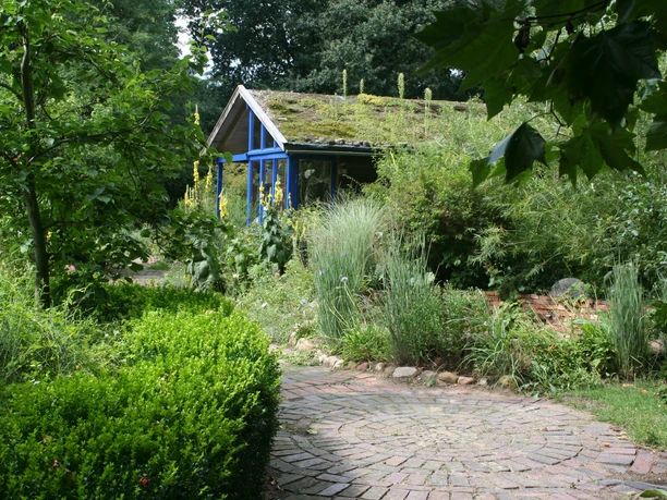 BUND Naturnaher Schau- und Lehrgarten Vielseitiger, naturnaher Lehrgarten mit geschwungenem Kiesweg, üppigem Grün und einem Moos-bedeckten Holzhaus.