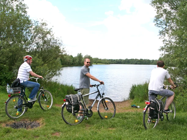 Radfahrer an der "Nienburger Rolle" Drei Radfahrer halten mit Blick auf einen ruhigen See und umgeben von grüner Vegetation inne.