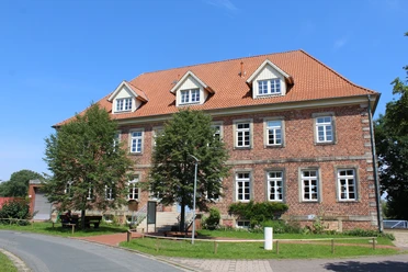 Amtshaus Westen mit Heimatstube Backsteingebäude mit roten Dachziegeln und drei Dachgauben, umgeben von Bäumen und einer Straße.