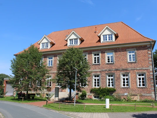 Amtshaus Westen mit Heimatstube Backsteingebäude mit roten Dachziegeln und drei Dachgauben, umgeben von Bäumen und einer Straße.