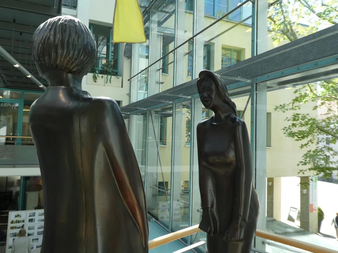 Zwei moderne Bronze-Skulpturen stehen sich gegenüber in einem lichtdurchfluteten Gebäudekomplex.