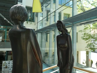 Skulptur Der erste Streit Zwei moderne Bronze-Skulpturen stehen sich gegenüber in einem lichtdurchfluteten Gebäudekomplex.