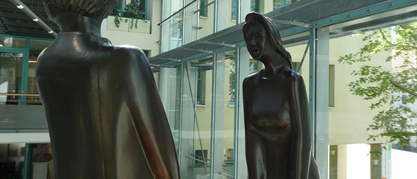 Skulptur Der erste Streit Zwei moderne Bronze-Skulpturen stehen sich gegenüber in einem lichtdurchfluteten Gebäudekomplex.