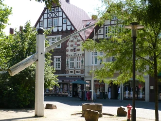 Skulptur eines überdimensionalen Nagels vor Fachwerkhäusern im Sonnenschein, umgeben von Bäumen.