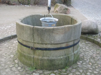 Runder steinerner Brunnen mit hängendem Eimer über gepflastertem Boden in parkähnlicher Umgebung.
