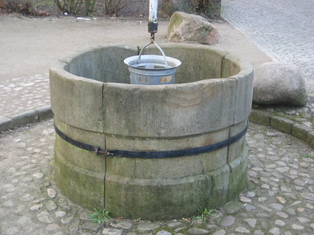 Skulptur Der Hungerbrunnen Runder steinerner Brunnen mit hängendem Eimer über gepflastertem Boden in parkähnlicher Umgebung.