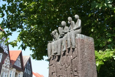 Skulptur von sitzenden Figuren auf einem hohen Sockel, umgeben von grünen Blättern und Fachwerkhäusern.