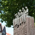 Skulptur von sitzenden Figuren auf einem hohen Sockel, umgeben von grünen Blättern und Fachwerkhäusern.