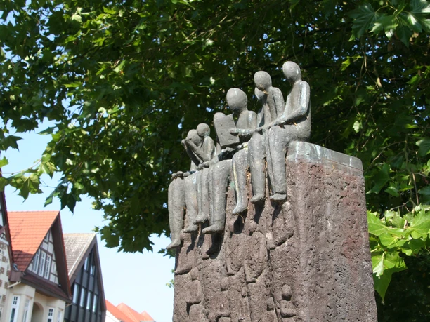 Skulptur "Der Vorleser" Skulptur von sitzenden Figuren auf einem hohen Sockel, umgeben von grünen Blättern und Fachwerkhäusern.
