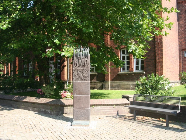 A bronze sculpture depicting figures reading, set against a backdrop of red brick buildings and green trees.