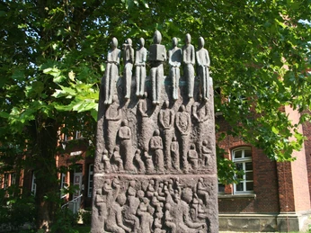 Bronzeskulptur von lesenden Figuren, im Schatten eines großen Baums vor einem Backsteingebäude.