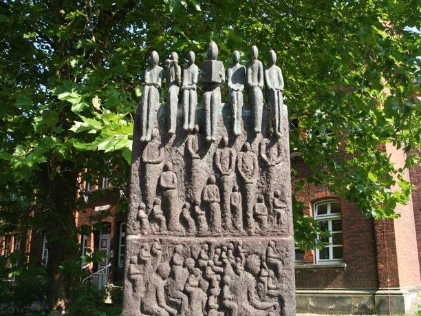 A bronze sculpture of figures reading, in the shade of a large tree in front of a brick building.