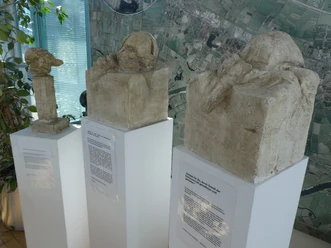 Skulptur Epitaph Drei abstrakte Skulpturen auf weißen Sockeln vor einer Karte zeigen unterschiedliche menschliche Kopfformen.