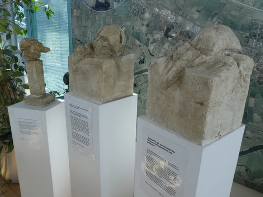 Skulptur Epitaph Drei abstrakte Skulpturen auf weißen Sockeln vor einer Karte zeigen unterschiedliche menschliche Kopfformen.