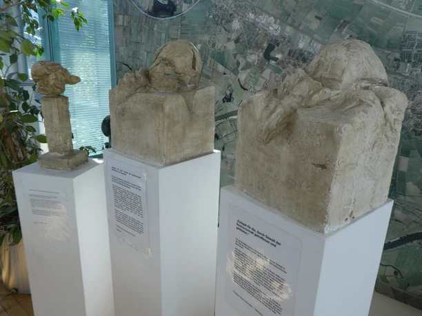 Skulptur Epitaph Drei abstrakte Skulpturen auf weißen Sockeln vor einer Karte zeigen unterschiedliche menschliche Kopfformen.