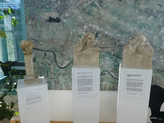 Drei stelenartige Skulpturen stehen vor einer großen, detaillierten Luftaufnahme einer Landschaft.