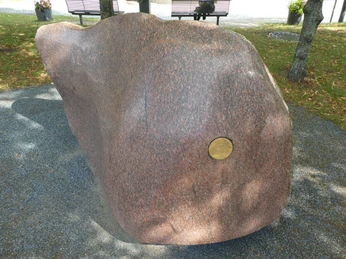 Rötlich brauner Granitblock mit eingelassener Gedenktafel, umgeben von Bäumen und Schotterweg.