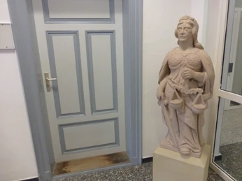 Eine steinerne Justitia-Skulptur mit Waage und Schwert steht drinnen neben einer grauen Tür.