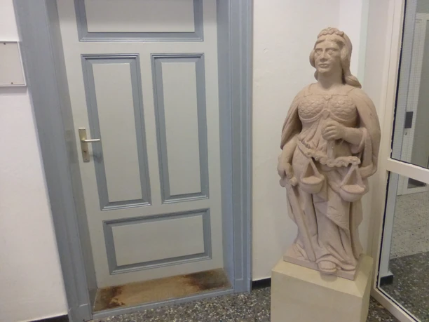 Skulptur "Jusitia" Eine steinerne Justitia-Skulptur mit Waage und Schwert steht drinnen neben einer grauen Tür.