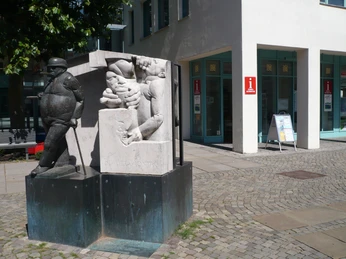 Skulptur "Zigarrenmacher" Die Skulptur "Zigarrenmacher" zeigt einen Handwerker, der Zigarren rollt, vor einem modernen Gebäude.