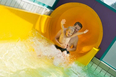 100 Meter lange Riesenrutsche Vater und Kind rutschen lachend aus einer großen gelben Wasserrutsche ins spritzende Becken.