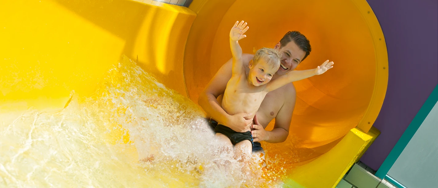 100 Meter lange Riesenrutsche Vater und Kind rutschen lachend aus einer großen gelben Wasserrutsche ins spritzende Becken.