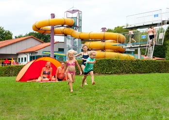 Hallen- und Freibad Wingst Kinder rennen lachend über die Liegewiese vor gelber Wasserrutsche im Hallen- und Freibad Wingst.