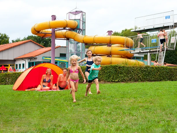 Hallen- und Freibad Wingst Kinder rennen lachend über die Liegewiese vor gelber Wasserrutsche im Hallen- und Freibad Wingst.