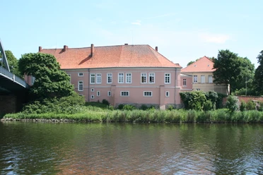 Schloß Hoya Historisches Schloß Hoya am Ufer eines Flusses, umgeben von Bäumen unter blauem Himmel.