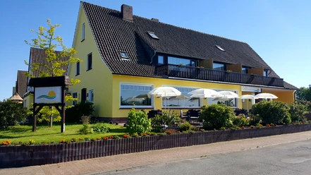 Café Sonnenschein Das Bild zeigt das Café und die Pension Sonnenschein. Im Vordergrund sind gemütliche Sitzgelegenheiten und drei weiße Sonnenschirme zu sehen. Im Hintergrund erstrahlt die Pension in einem warmen Gelbton.