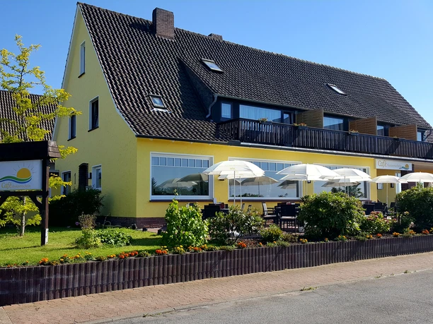 Café Sonnenschein Das Bild zeigt das Café und die Pension Sonnenschein. Im Vordergrund sind gemütliche Sitzgelegenheiten und drei weiße Sonnenschirme zu sehen. Im Hintergrund erstrahlt die Pension in einem warmen Gelbton.