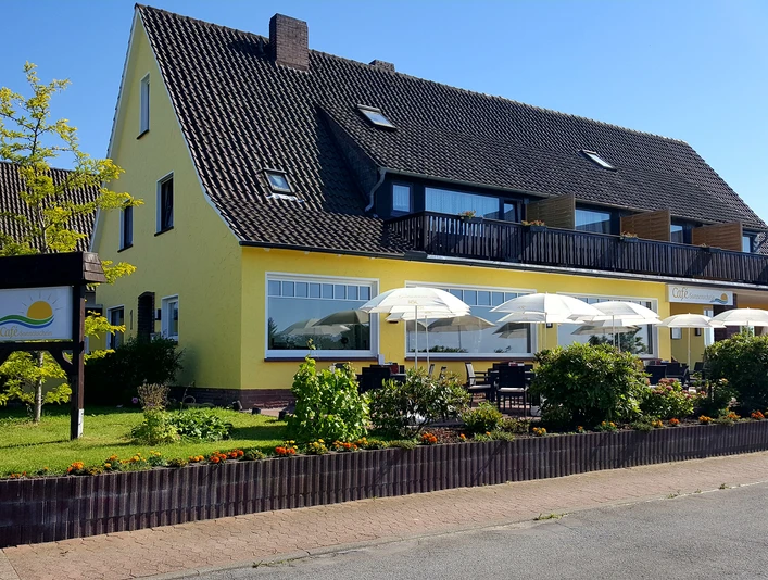 Café Sonnenschein Das Bild zeigt das Café und die Pension Sonnenschein. Im Vordergrund sind gemütliche Sitzgelegenheiten und drei weiße Sonnenschirme zu sehen. Im Hintergrund erstrahlt die Pension in einem warmen Gelbton.