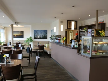 Gastraum des Café Sonnenschein Das Bild zeigt die gemütliche Einrichtung des Cafés, das in warmen Braun- und Naturtönen gestaltet ist und eine behagliche Atmosphäre bietet. Links im Bild sieht man eine Theke, die mit einer verlockenden Auswahl an Kuchen zum Genießen einlädt.