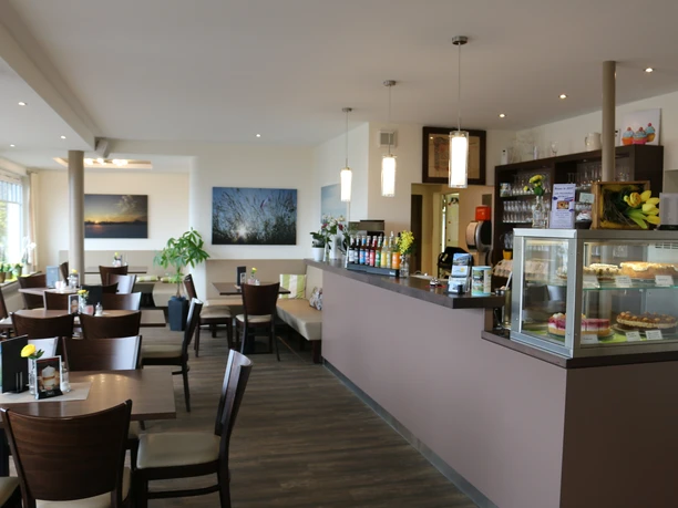 Gastraum des Café Sonnenschein Das Bild zeigt die gemütliche Einrichtung des Cafés, das in warmen Braun- und Naturtönen gestaltet ist und eine behagliche Atmosphäre bietet. Links im Bild sieht man eine Theke, die mit einer verlockenden Auswahl an Kuchen zum Genießen einlädt.