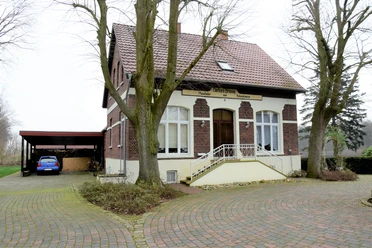 Moorlädchen Backsteinhaus mit hellem Giebel, breiter Treppe und Carport, umgeben von Bäumen und Pflasterweg.