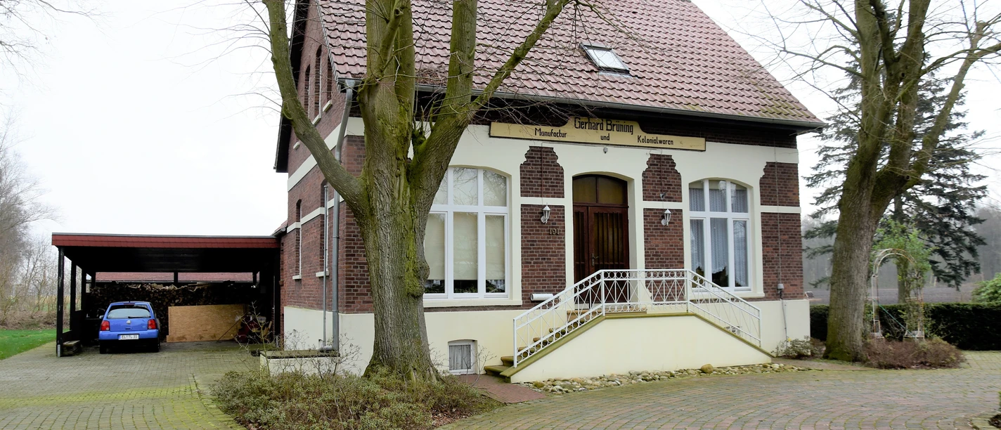 Moorlädchen Backsteinhaus mit hellem Giebel, breiter Treppe und Carport, umgeben von Bäumen und Pflasterweg.