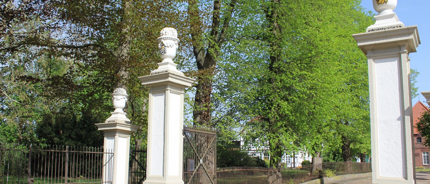 Eingang des historischen Ritterguts von Behr, gesäumt von Säulen mit dekorativen Vasen in ländlicher Umgebung.